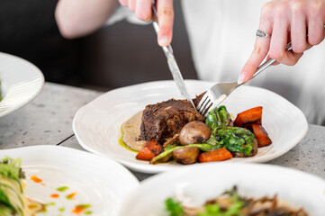 Close-up of a white plate with cooked meat, fresh vegetables, and garnish, being cut with a knife and fork on a dining table. Ideal for concepts of fine dining and culinary presentation.
