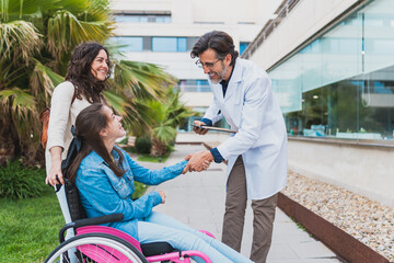 Doctor greeting young woman in wheelchair with caregiver outdoors - Smiling doctor greets young...