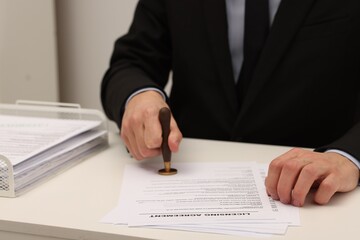 Man stamping licensing agreement document at white table indoors, closeup