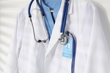 Doctor's coat with stethoscope and badge on hanger against white wall, closeup