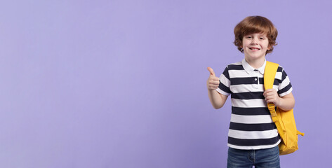 Naklejka premium Cute schoolboy with backpack showing thumbs up on violet background, space for text. Banner