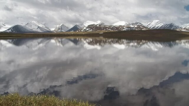 This 4K footage captures a lake in Arabel Plateau, Kyrgyzstan, with small currents rippling the water, reflecting snowy mountain peaks and clouds in a serene landscape