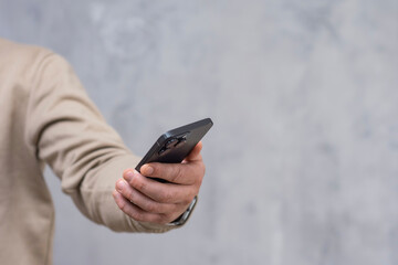A man in a brown sweatshirt holds a smartphone against a gray concrete wall. Modern technology and use of mobile devices.