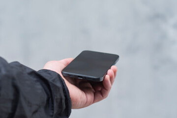 A man in a black jacket holding a smartphone against a gray concrete wall. Modern technology and the use of mobile devices.