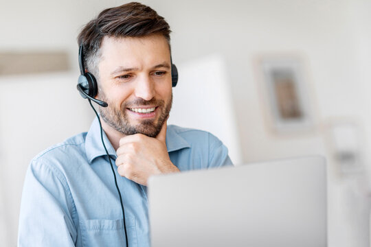 Hotline Support Service. Handsome male call center operator in headset working with laptop in modern office, looking at computer screen, free space