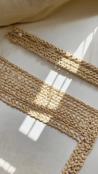 Handmade woven raffia placemat and braided trim detail with shadows on white linen fabric, showcasing natural fibers, texture and craftsmanship.