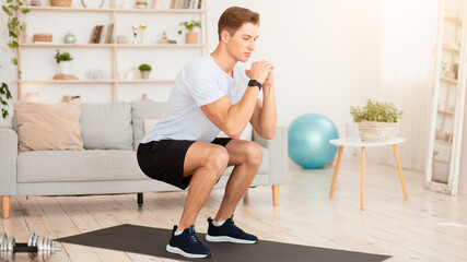 Obraz premium Online coach for home workout. Handsome young man in sportswear with fitness tracker, squats and looks at laptop in living room interior, free space