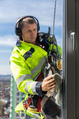 Worker in High Visibility Gear Cleans Window on High-Rise Building with Safety Gear, Blue Sky Background - A Scene of Precision and Skill in Window Cleaning Profession
