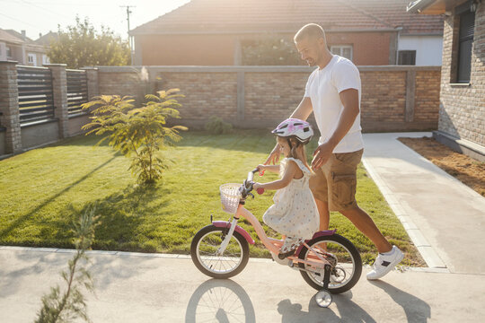 Young supportive father is teaching his little daughter how to ride bicycle while having fun in backyard.