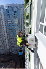 A Brave Worker Maintains Air Conditioning Units on a High-Rise Building, Showcasing Expertise in Extreme Heights and Ensuring Comfort for the Residents Below, Against a Clear Blue Sky