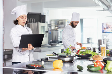 Dedicated chef using a laptop for research while cooking gourmet dishes, in a commercial kitchen balances technology and tradition. Expert chopping vegetables and preparing ingredients.