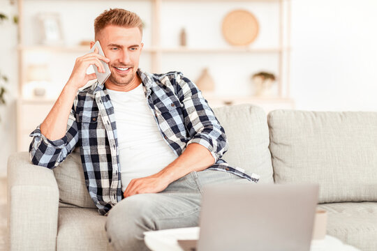 Phone Call. Portrait of confident adult man talking on cellphone, sitting on the couch in living room, looking at computer. Communication, Rest And Relax Concept. Copy space. Guy having conversation