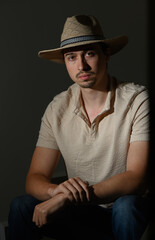 Portrait of a young man in a straw hat seated in dim lighting, reflecting thoughtful contemplation in a stylish setting © Михаил Шорохов