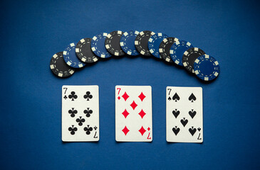 Poker chips and playing cards are arranged neatly on a blue table. The cards show three sevens,...
