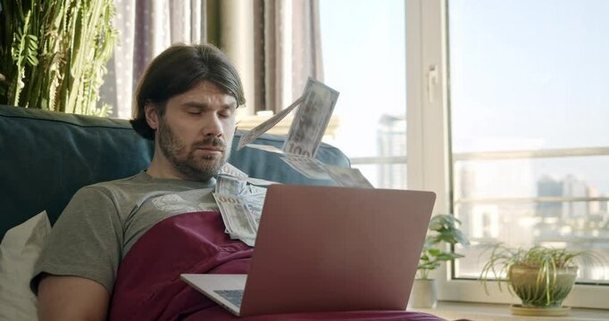 Freelancer man making money online using laptop, lying on bed, earn money from the comfort of bed. Cash flow. Money rain flow through the laptop. Dollar bills flying in the investor face