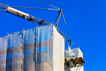 Tall building under construction is wrapped in white scaffolding and protective netting, with a crane looming overhead against a clear blue sky. Scene captures the essence of urban development