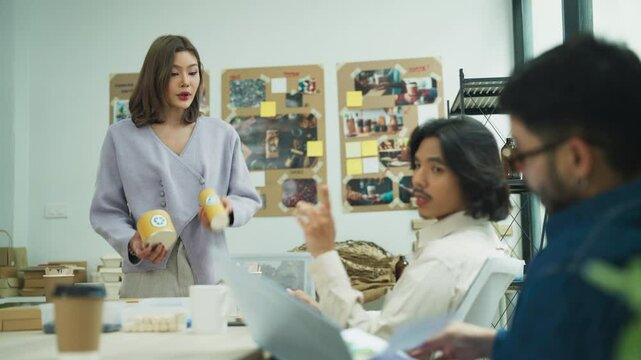 Young Asian businesswoman confidently presenting eco-friendly packaging products to diverse colleagues during a collaborative team discussion, emphasizing sustainability and creative solutions.