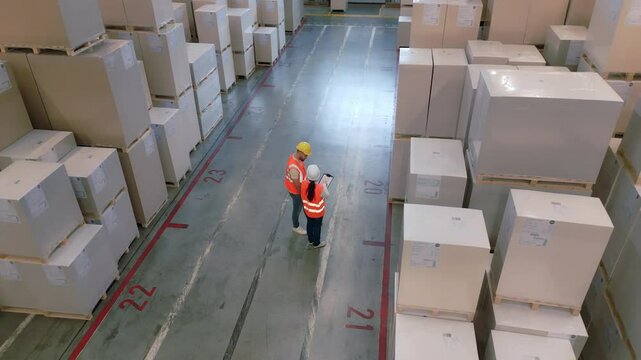 A bird's-eye view of a bustling warehouse reveals two workers in orange vests and hard hats navigating between towering stacks of white crates, discussing logistics and distribution strategies.