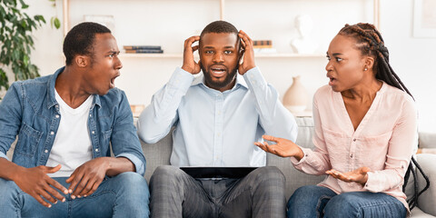 Shocked black marital counselor sitting between arguing spouses during therapy session, covering...