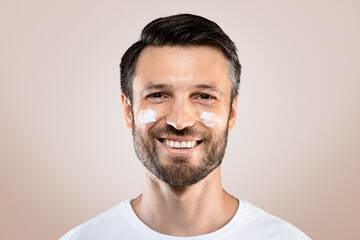 Obraz premium Morning routine. Young happy man with face cream on his cheeks, grey studio background, copy space. Handsome man with face cream or lotion smiling at camera, beauty products for men, male cosmetology