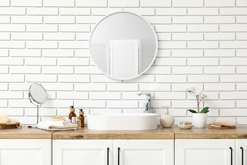 Sink, bottles of cosmetic products on chest of drawers and mirror on white brick wall in bathroom