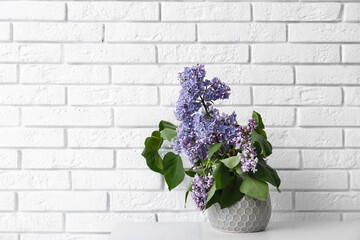 Vase with lilacs branches on shelf near white brick wall in room