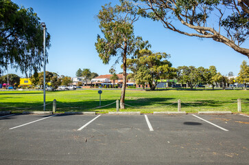 Empty parking spaces in an open-air parking lot with marked bays facing a green park and a distant suburban commercial street. Car park at a parkland or near public community spaces in Australia.