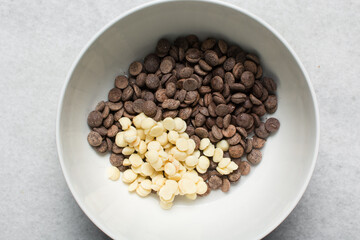 Overhead view of dark chocolate chips and white chocolate chips in a white bowl, top view of chocolate chips on a white background