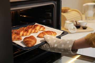 Woman taking baking sheet with tasty croissants out of oven indoors, closeup