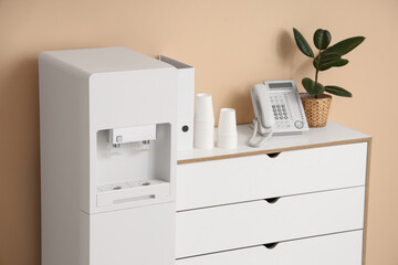 Modern water cooler and chest of drawers near beige wall