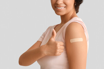 Young woman with medical patch showing thumb-up after vaccination on light background, closeup