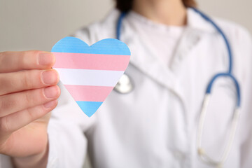 Female doctor with paper heart in colors of transgender flag on light background, closeup