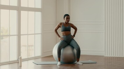 A young African American woman engages in a core workout while seated on a stability ball. The bright and airy indoor space enhances her focus on fitness and healthy living.