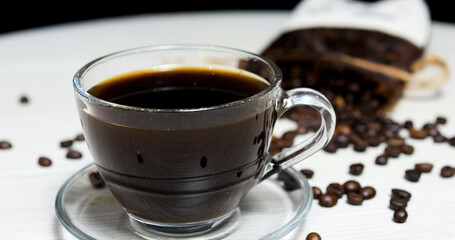 Steaming black coffee sits in a glass cup on a white table, surrounded by roasted coffee beans spilling from a burlap sack, creating a cozy and inviting scene for coffee lovers