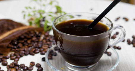 Steaming cup of black coffee sits on a saucer with a black stirrer, surrounded by roasted coffee beans and a green coffee plant on a light wooden table, creating a cozy and inviting atmosphere