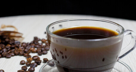 Transparent glass cup filled with steaming black coffee sits on a white table alongside scattered roasted coffee beans and a small paper bag, creating a cozy and inviting coffee break scene