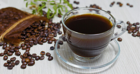 Transparent coffee cup filled with dark, aromatic coffee sits on a glass saucer, surrounded by scattered roasted coffee beans and a decorative burlap bag
