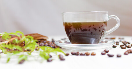 Transparent glass cup filled with steaming coffee sits on a saucer on a white table, surrounded by scattered roasted coffee beans and a small green plant, creating a cozy and inviting atmosphere
