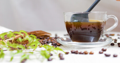 Transparent coffee cup sits on a saucer with a spoon stirring the dark coffee, surrounded by roasted coffee beans and a green plant on a white wooden table