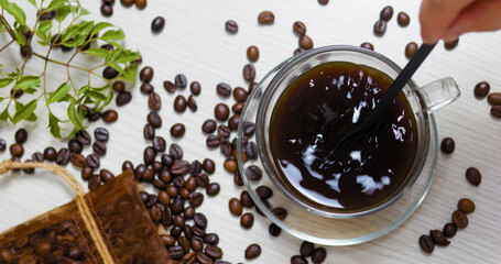 Barista is stirring a transparent cup of coffee with a black spoon, surrounded by roasted coffee beans, a small green plant and a burlap sack, creating a cozy and aromatic coffee break scene