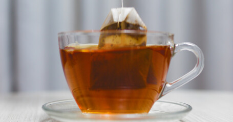 Tea bag steeping in a glass cup of hot water, placed on a saucer, sits on a table against a softly blurred background, creating a cozy and comforting scene