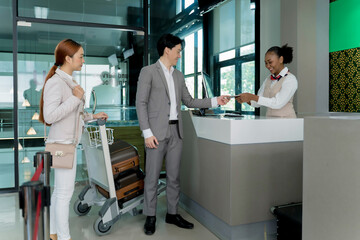 Business travelers handing over passports and tickets to airline staff at airport check-in counter, ready for departure with luggage on trolley and friendly service