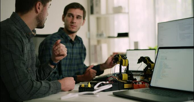 Software engineers work side-by-side in a tech lab, testing robotic arms through programmed code displayed on laptops and monitors.