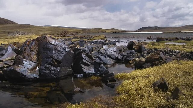 Footage captures the rugged beauty of Arabel Plateau, Kyrgyzstan, showcasing volcanic rocks along a rocky shoreline with patches of vegetation bordering a body of water.