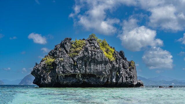 A rocky, lonely little island in a turquoise ocean. There is green vegetation on the steep, furrowed slopes. Blue sky, clouds. Philippines. Palawan. Bacuit bay