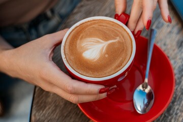 Coffee Latte Art Red Cup Hands Cafe. Close-up of a hand holding a red mug of coffee with latte art, a spoon, and a red saucer on a wooden table.