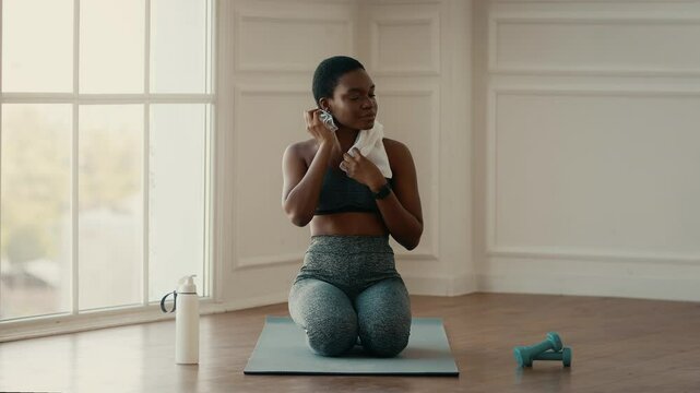A young African American woman takes a breather after an intense workout session at home. She kneels on a yoga mat, towel in hand, showcasing her commitment to a healthy lifestyle.