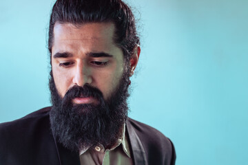 Close-up portrait of man with full, dark beard and hair pulled back in topknot. wearing dark blazer over light-colored shirt, his expression is pensive, his gaze directed downwards.