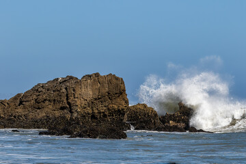 Giant wave breaking against large rock just offshore Malibu, California. Spray flying high in the air; blue sky in the distance. 
