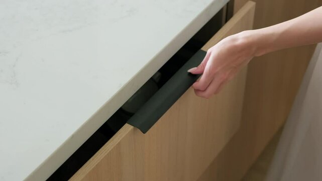 Kitchen storage. Young woman places a clear glass teapot with a wooden lid into a modern kitchen drawer. Minimalist wooden cabinet, modern furniture